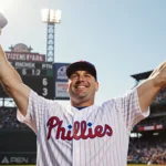 Rob Thomson triumphantly raises arms behind home plate with a beaming smile and a scoreboard reading Phillies 4 opponent 3