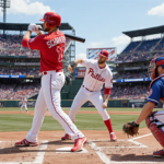 Kyle Schwarber at home plate with bat slung over shoulder as pitcher winds up to throw a fastball and Phillies fans cheer.