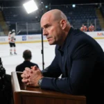 Rick Tocchet head coach delivering press conference with frustrated under spotlight and blurred Flyers practice rink