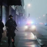 Two injured officers stand beside a shattered storefront window with flashing police lights and foggy street