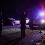 Teen suspect raising arms with police car flashing lights and dark Gloucester street during nighttime arrest