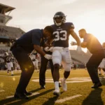 Quinshon Judkins being helped off the field with an ice pack on his injured leg and teammates watching at sunset