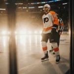 Rasmus Ristolainen stands ready on the Flyers’ ice with Cam York beside him lights reflecting off glass.