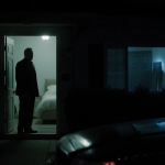 Silhouetted home at night with front door ajar and glowing hallway behind dark bedroom faint police car reflection foreboding