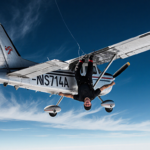 Skydiver hanging upside-down from Cessna Caravan stabilizer with parachute tangled on tail and deep blue sky.