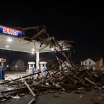 Sunoco gas station roof collapsing at night with twisted metal beams and debris around pumps.