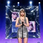 Taylor Swift performing on stage with spotlight and metallic mirror reflecting album art and glowing blue-purple backdrop