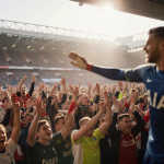 Fans cheering with colors in Tottenham Hotspur Stadium and blurred Liverpool goalkeeper and warm light from large windows