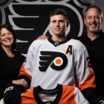 Ty Murchison stands triumphant in front of a Philadelphia Flyers logo with his parents beside him and a sunrise behind