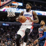 Tyrese Maxey driving to the basket with Paul George watching from the bench and Philadelphia skyline visible behind arena.