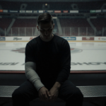 Tyson Foerster sits on a bench with his injured arm bandaged and locker room near a glass wall with a blurred Flyers ice show