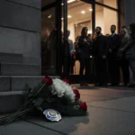 Mourners gather outside University of Delaware Bob Carpenter Center with white floral arrangement and police badge on steps