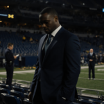 Sherrone Moore looks down in a lit Michigan football stadium with scattered seats and a blurred administrator in background.