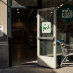Abandoned Amazon Go storefront opens with a crooked sign and faded Fresh logo with door ajar and cart beside Whole Foods box