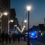 AI surveillance cameras watch East Lansdowne street at dusk with people walking and police lights flashing