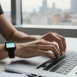 Young adult types on laptop with Bee wearable device glowing on wrist and city lights through window behind