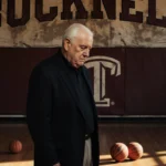 Bill Courtney stands before faded Bucknell banner with bowed head and scattered basketballs showing coaching legacy