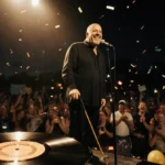 Billy Joel stands at microphone with a cane while a vinyl record and confetti light up the stage.