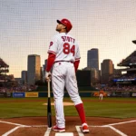 Bryson Stott at home plate with bat on leg looking up at mound under lights of Citizens Bank Park during sunset