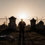 Security guard stands at cemetery fence with setting sun casting long shadows across overgrown graves