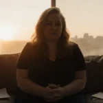 Colleen Hoover sits contemplatively with clasped hands and books nearby as sunset glows through window