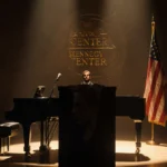 Grand piano sits in dimly lit ornate concert hall with a golden stage glow and a tattered flag showing Kennedy Center logo