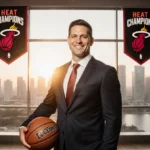 Erik Spoelstra stands proudly with basketball and Heat championship banners with Miami skyline glowing behind him