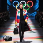 Man struts down fashion runway with vibrant graphics and Olympic rings in background while designer coat stands out with gem