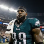Brandon Graham standing with Eagles jersey draped over shoulder facing opponent on football field with stadium lights crowd
