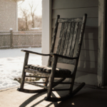 Weathered rocking chair angled on porch with faint human shadow and snow-covered backyard in cold light