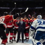Philadelphia Flyers players huddle with coach while fans wave towels and Lightning goalie guards net with sticks raised