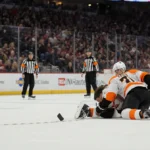 Travis Konecny lies on ice clutching his knee with teammates helping and puck resting nearby