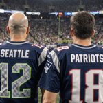 Two football coaches stand on a football field with Super Bowl LX jerseys showing their names and a stadium crowd behind.