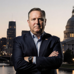 Governor Tim Walz standing at dusk with Minneapolis skyline and Capitol behind him arms crossed and determined.