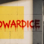 Wall displays yellow graffiti with red letters COWARDICE under morning light and open school door.