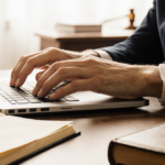 Hand typing on laptop with warm golden light and a leather book beside blurred courtroom background hinting tension