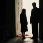 Woman standing at home entrance with subtle blood sheen on floor and faint glow inside.