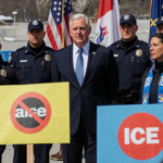 Vice President Vance stands at venue while ICE agents in uniforms hide behind him and protestors hold anti‑ICE signs.