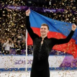 Ilia Malinin celebrates victory on ice with arms raised and confetti falling while flag waves behind him