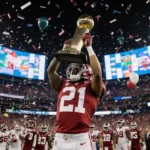 Indiana football player lifts championship trophy with confetti and stadium lights while screens show semifinal scores.