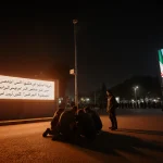 Protesters huddle around smuggled Starlink terminal with LED billboard showing Farsi message and Raisi portrait in background