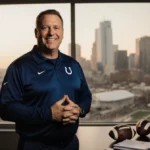 Jim Bob Cooter stands with clasped hands and confident smile before the Indianapolis skyline with footballs and playbooks on