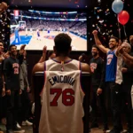 Joel Embiid sitting with crossed arms while giant screen shows game highlights in dim Madison Square Garden suite