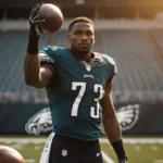 Jordan Mailata standing behind the Eagles offense line holding a football aloft with warm golden lighting and empty seats