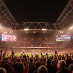 Excited football fans cheer in packed stadium with team colors and giant screens showing match highlights