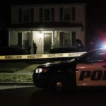 Police officers guard a suburban home at night with crime scene tape and cruiser lights illuminating the porch