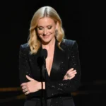 Nikki Glaser stands alone on the Golden Globes stage with dropped microphone and crossed arms while golden light highlights h