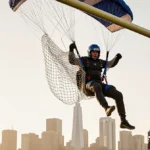 Parachutist tangled in netting behind a goal post with dynamic golden lighting and a blurred Fort Worth backdrop.