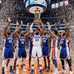 Joel Embiid basketball holding championship trophy with confetti exploding and Spectrum Arena seats orange blue silver