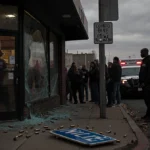 Concerned citizens gather behind police car and ambulance with broken storefront and shattered street sign